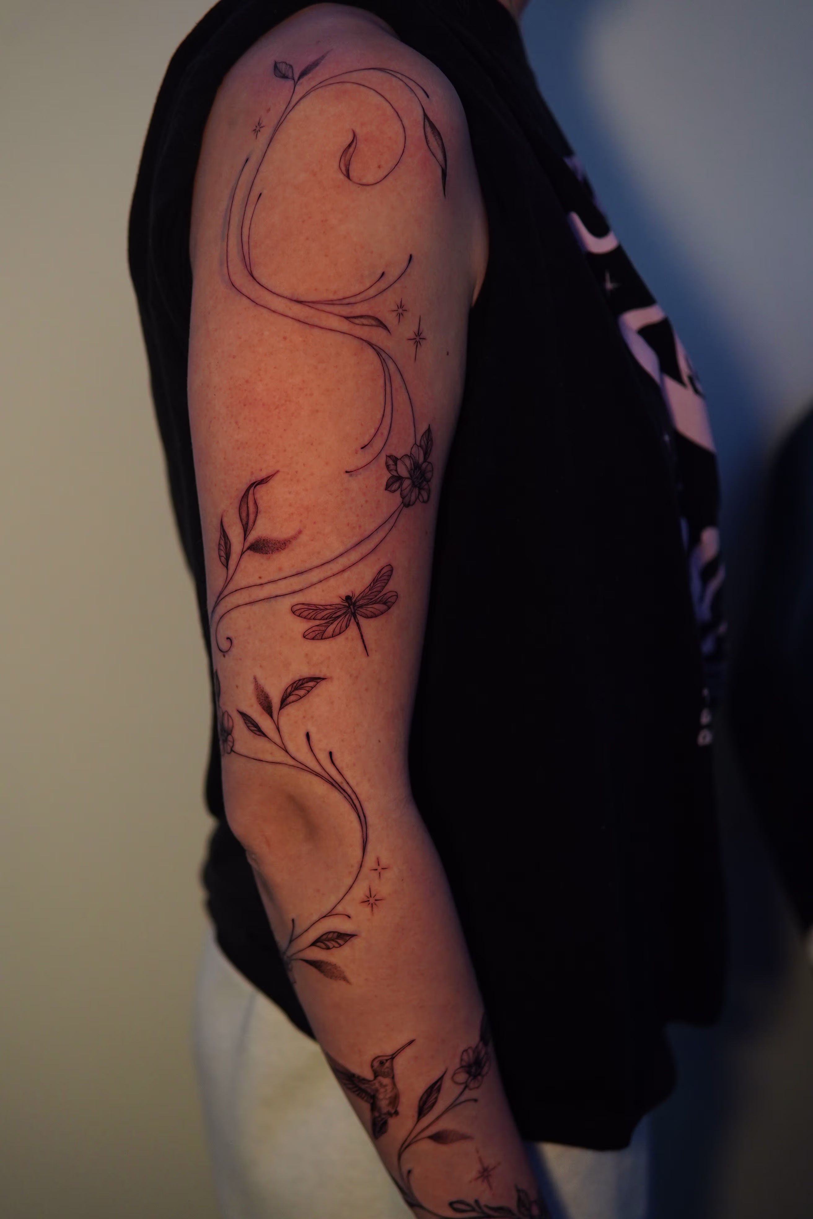 Wrap around fine-line vine with leaves, flowers, sparkles, a hummingbird and dragonfly, starting from the right wrist up to the shoulder.